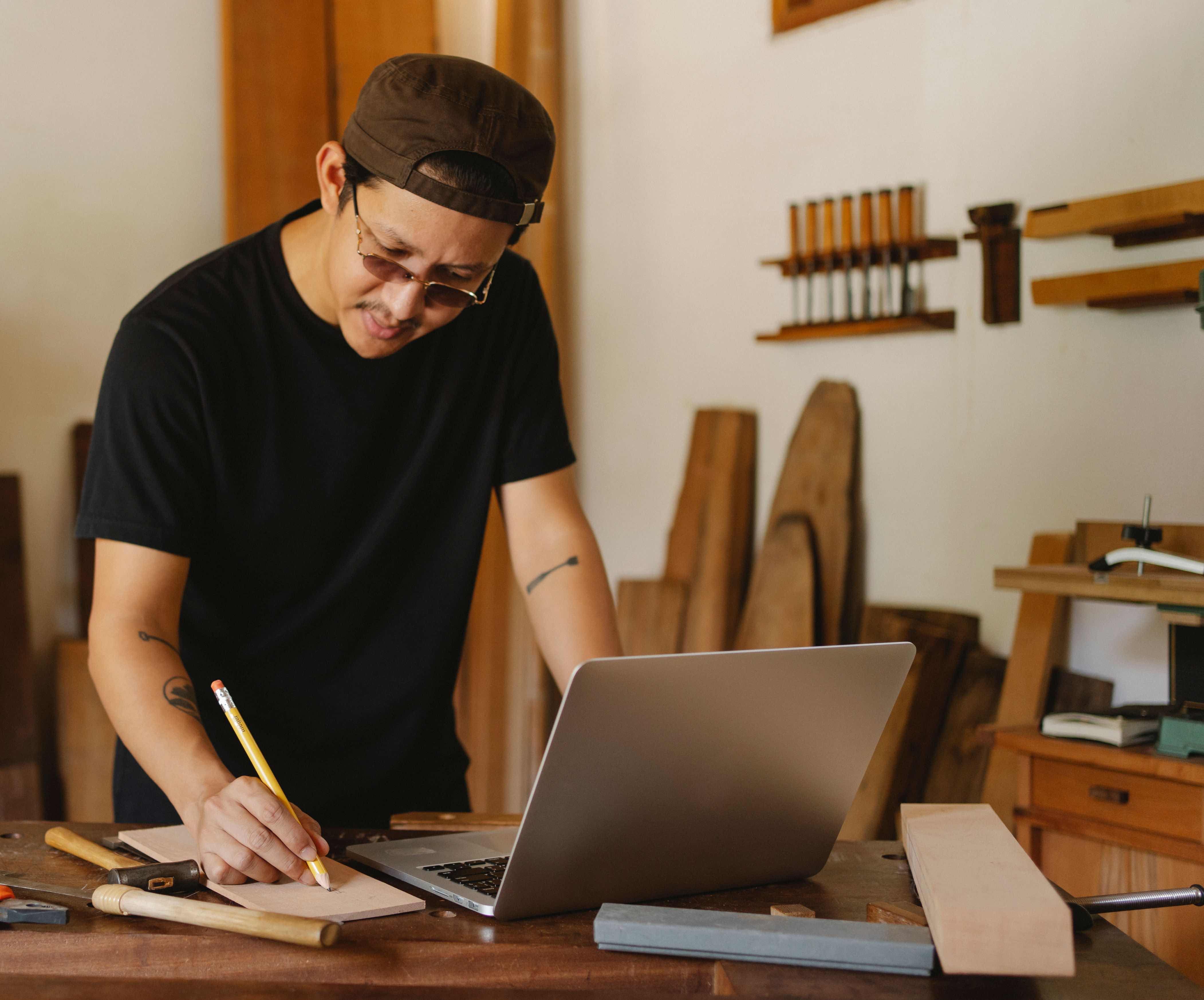 A carpenter reviewing notes beside a laptop and hand tools in a workshop
